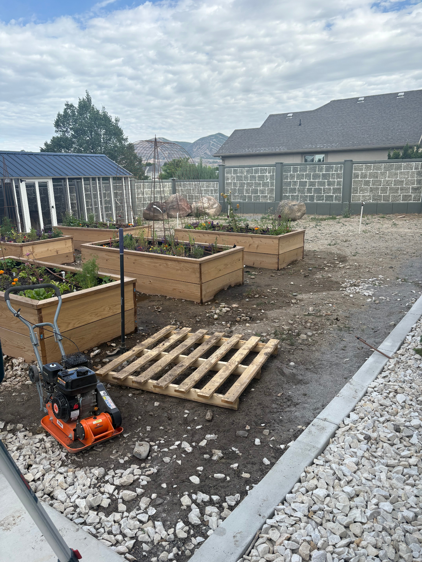 Before photo of a Utah backyard mid-build: dirt ground with wooden raised garden beds under construction, a compactor and pallet on site, and a greenhouse to the left.