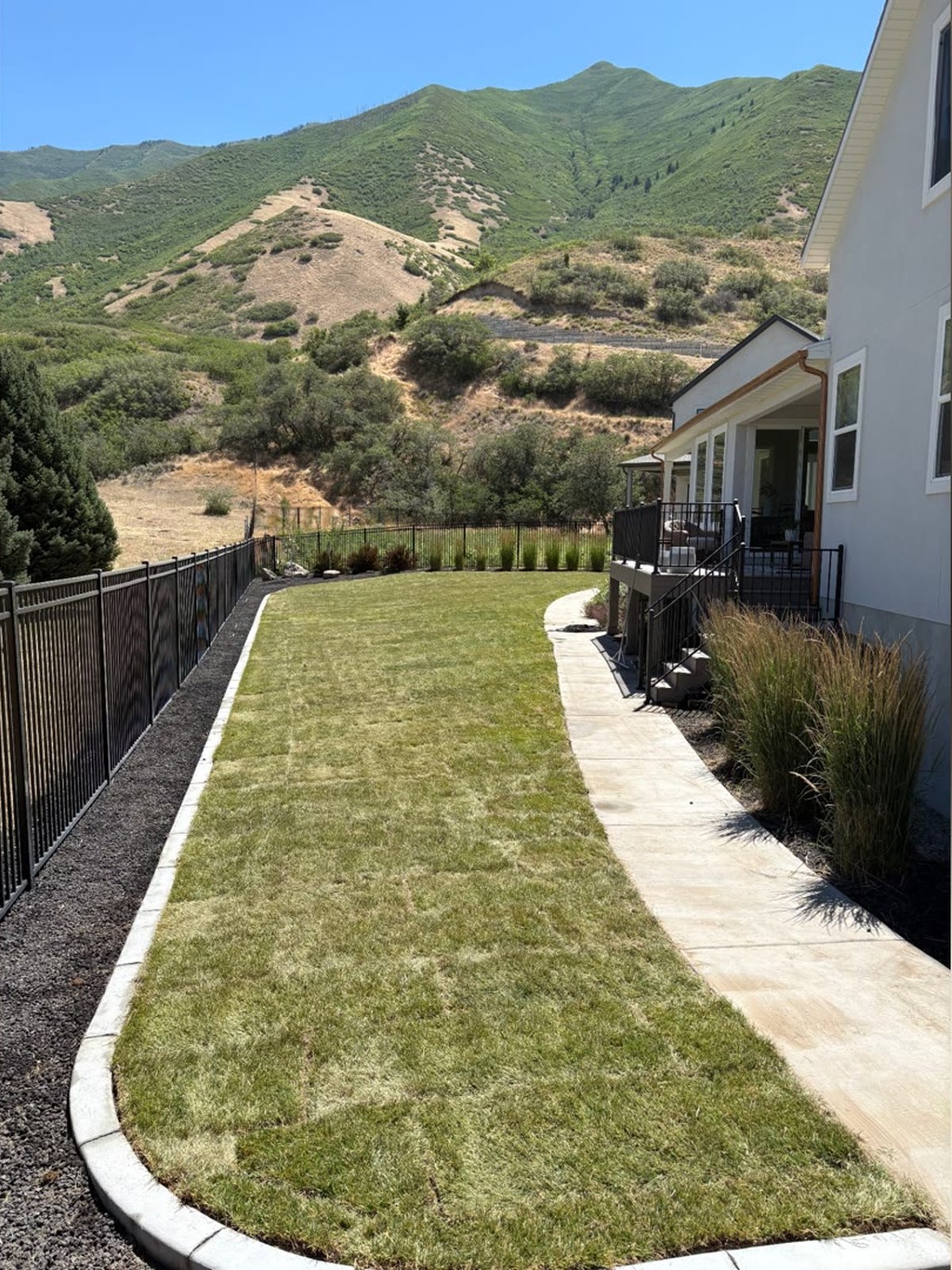 After photo of the same Utah yard: lush green sod lawn edged with clean white concrete curbing, mulched flower beds, and Wasatch mountain views — landscaping completed by Blue Wing Construction and Landscape.